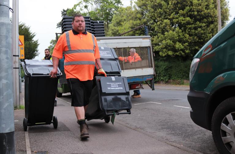 A binman in orange high vis wheels two bins down the street to deliver them to households