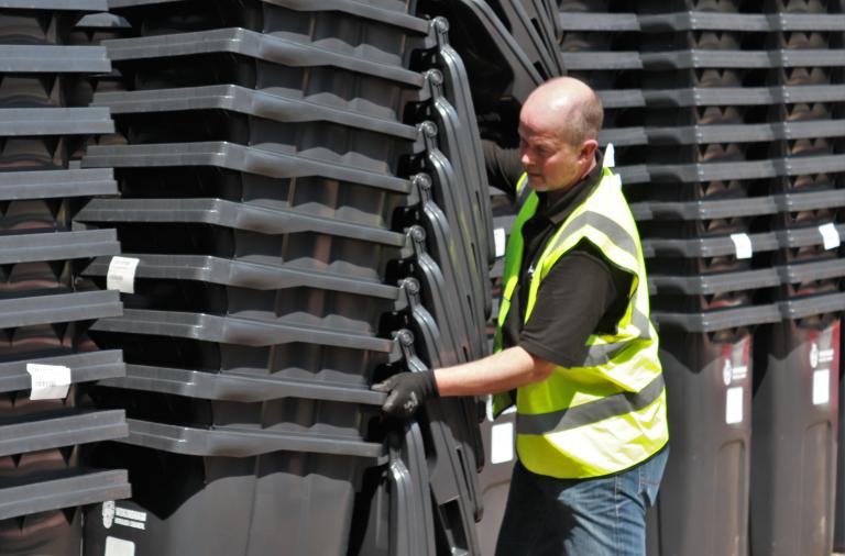 A man in a high vis jacket stacks a column of black bins next to loads of other columns of stacked black bins