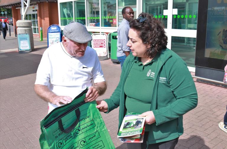 A smiling woman in a council fleece hands a green recycling bag to an older man outside Asda in Lower Earley