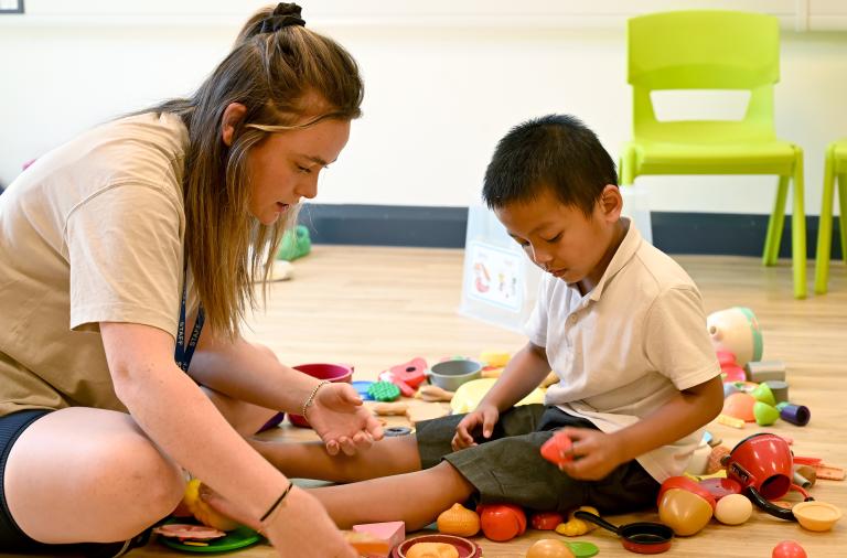 A boy sits on the floor with an adult surrounded by role play food