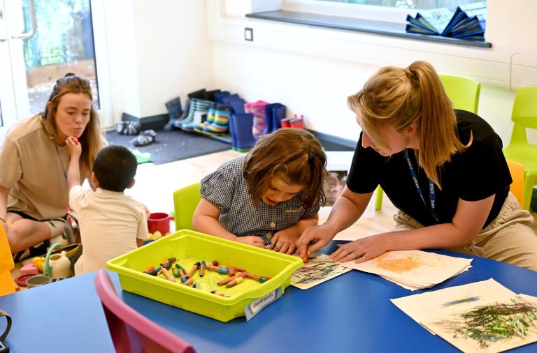 A child sits colouring at a table with an adult, in the background a boy pretends a crayon is lipstick on another adult