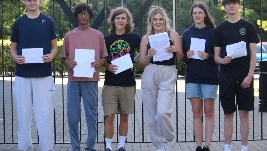 Six students stand in a row smiling and holding up their A level results