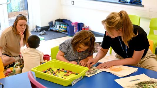 A child sits colouring at a table with an adult, in the background a boy pretends a crayon is lipstick on another adult